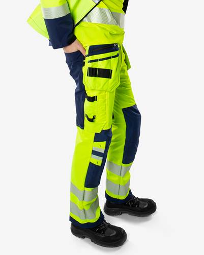 Eine Person trägt Sicherheitsausrüstung mit hoher Sichtbarkeit, einschließlich heller gelber Hose mit reflektierenden Streifen und Taschen, und steht vor einem weißen Hintergrund.
