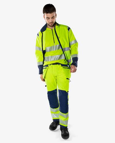 Eine Person, die Arbeitskleidung mit Warnfarben und reflektierenden Streifen trägt, einschließlich einer gelben Jacke und Hose mit blauen Akzenten, die vor einem neutralen Hintergrund steht.