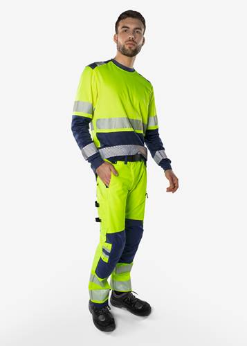 Eine Person in Warnschutzkleidung mit reflektierenden Streifen steht mit der Hand in der Tasche vor einem einfarbigen Hintergrund.