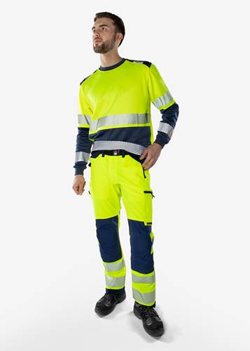 Eine Person, die eine neongelbe und marineblau-farbene Warnschutzkleidung trägt, bestehend aus Jacke, Hose und Schuhen, vor einem neutralen Hintergrund.