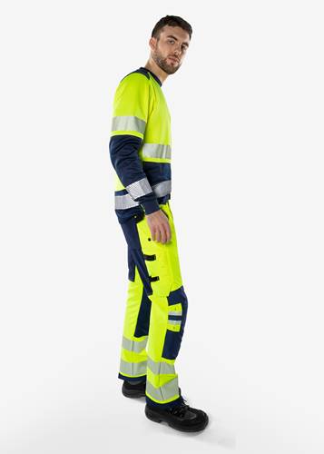 Eine Person in Sicherheitskleidung mit Signalwirkung, einschließlich einer neongelben Jacke und Hose mit reflektierenden Streifen, in einer Seitenansicht stehend.