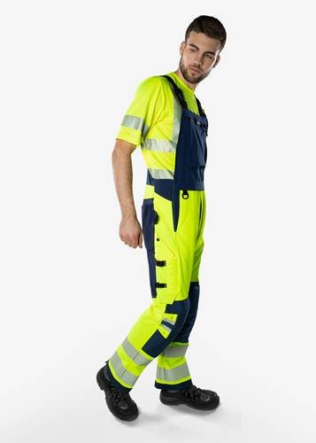 Eine Person, die einen Arbeitsanzug in Warnfarben mit reflektierenden Streifen trägt und vor einem einfarbigen Hintergrund posiert.