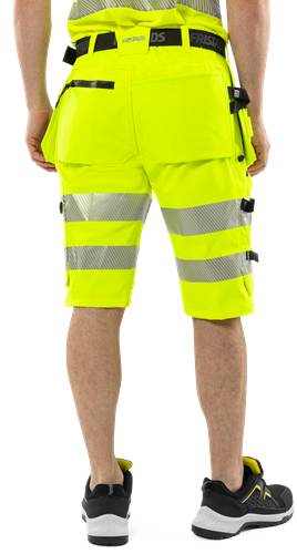 Eine Person, die in leuchtend gelben Arbeitsshorts mit reflektierenden Streifen vor einem schwarzen Hintergrund steht.