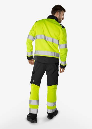 Eine Person, die eine Warnschutzjacke und -hose in knalligem Limettgrün mit reflektierenden Streifen trägt und wegblickend steht.