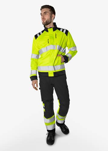 Eine Person, die eine hochsichtbare gelb-schwarze Sicherheitsjacke und -hose mit reflektierenden Streifen trägt und selbstbewusst einen Helm in der Hand hält.
