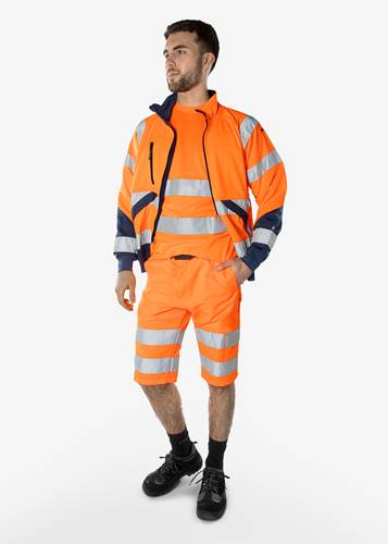 Eine Person, die leuchtend orangefarbene Arbeitskleidung mit hoher Sichtbarkeit und silbernen Reflektionsstreifen trägt, steht vor einem einfarbigen weißen Hintergrund.