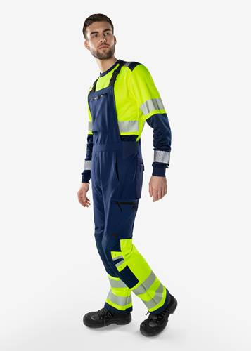Eine Person trägt einen Arbeitsschutzanzug in Marineblau und leuchtendem Gelb mit reflektierenden Streifen, die vor einem schlichten weißen Hintergrund steht.