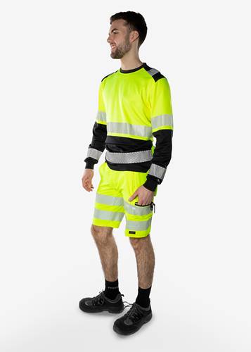 Eine Person trägt leuchtend gelbe Warnschutzkleidung mit Reflektorstreifen und steht vor einem neutralen Hintergrund, leicht nach rechts gewandt.
