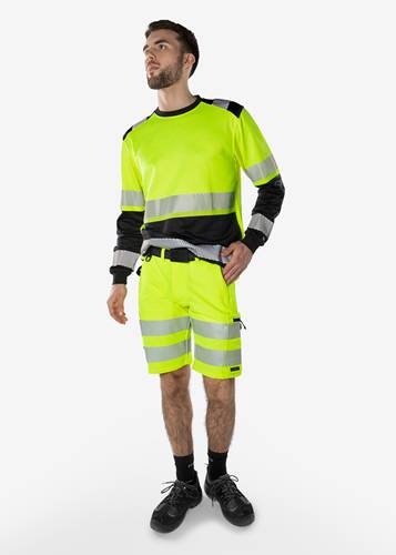 Eine Person in einer leuchtend gelben Sicherheitskleidung mit reflektierenden Streifen, bestehend aus einem langärmeligen Hemd und einer Shorts, vor einem einfarbigen Hintergrund.