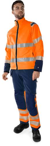 Eine Person, die eine leuchtend orangefarbene Warnschutzjacke mit reflektierenden Streifen und eine dunkelblaue Hose trägt, steht vor einem schwarzen Hintergrund.