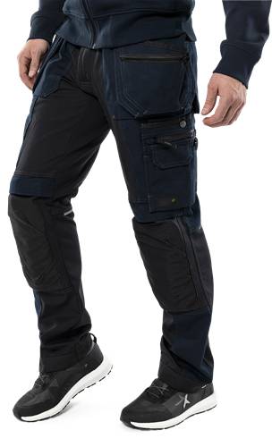 Person trägt strapazierfähige, dunkel gefärbte Arbeitshose mit mehreren Taschen, verstärkten Knien und hält etwas in der linken Hand.