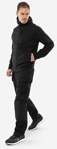 Eine Person, die eine schwarze Jacke und Hose trägt, mit der Hand teilweise in der Tasche. Die Kleidung wirkt lässig und funktional.