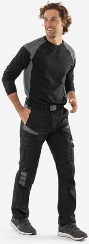 Eine Person mit schwarzer Cargohose und einem schwarzen und grauen Langarmshirt stützt eine Hand in der Tasche, vor einem neutralen Hintergrund stehend.