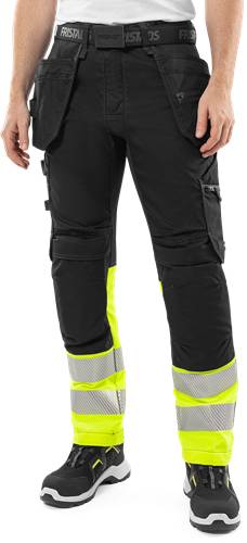 Person trägt schwarze Arbeitshose mit leuchtend gelben Reflektionsstreifen und mehreren Taschen vor schwarzem Hintergrund.
