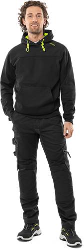 Eine Person in einem schwarzen Kapuzenpullover und Cargohose, mit der linken Hand teilweise in der Tasche, vor einem schlichten Hintergrund.