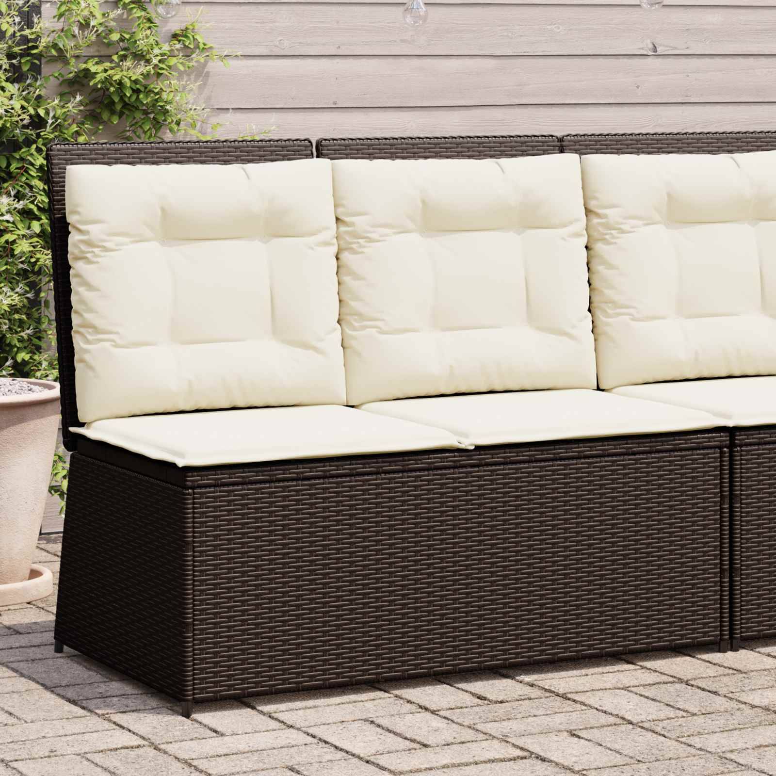 Ein brauner Geflecht-Terrassensofa mit cremefarbenen Kissen steht auf einer Ziegelsteinterrasse, an einer hellgrauen Holzwand mit grünen Pflanzen an der Seite platziert.