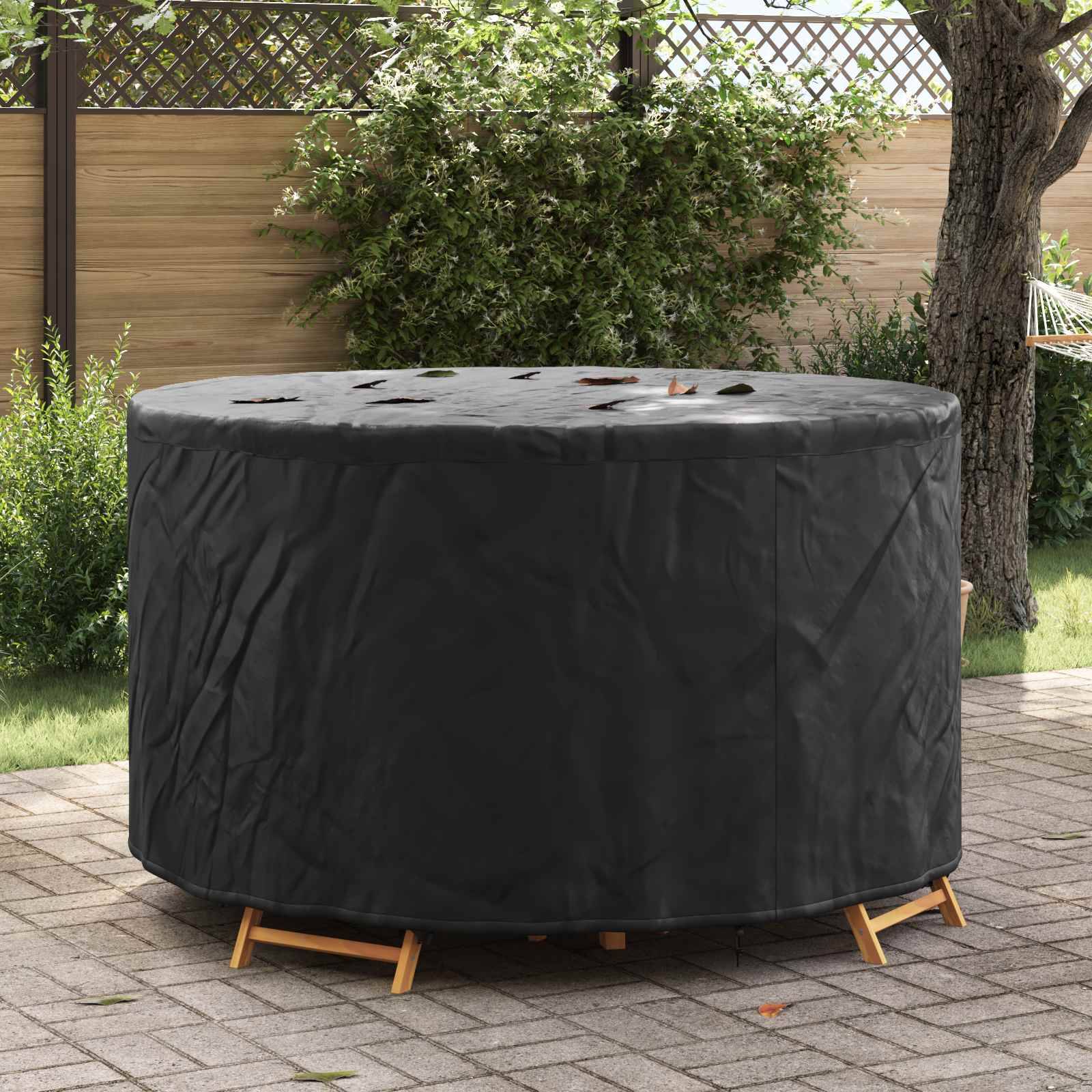 Eine runde Outdoor-Gartenmöbelgruppe, die mit einer schwarzen Schutzhülle bedeckt ist, auf einer mit Natursteinen gepflasterten Fläche in der Nähe eines Holzzauns und Grünpflanzen.