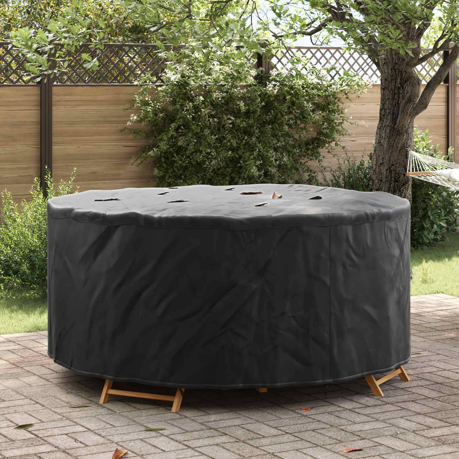 Ein runder Outdoor-Tisch, der mit einem schwarzen Schutzüberwurf bedeckt ist, auf einer Terrasse, umgeben von Bäumen und einem Holzzaun im Hintergrund.