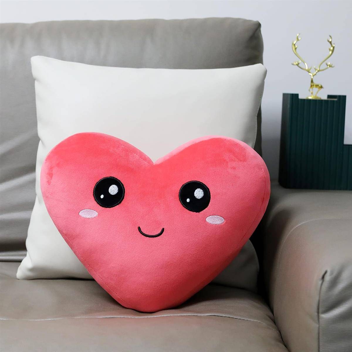 Rosafarbenes herzförmiges Plüschkissen mit einem süßen lächelnden Gesicht, das auf einem Sofa vor einem weißen Kissen ruht.