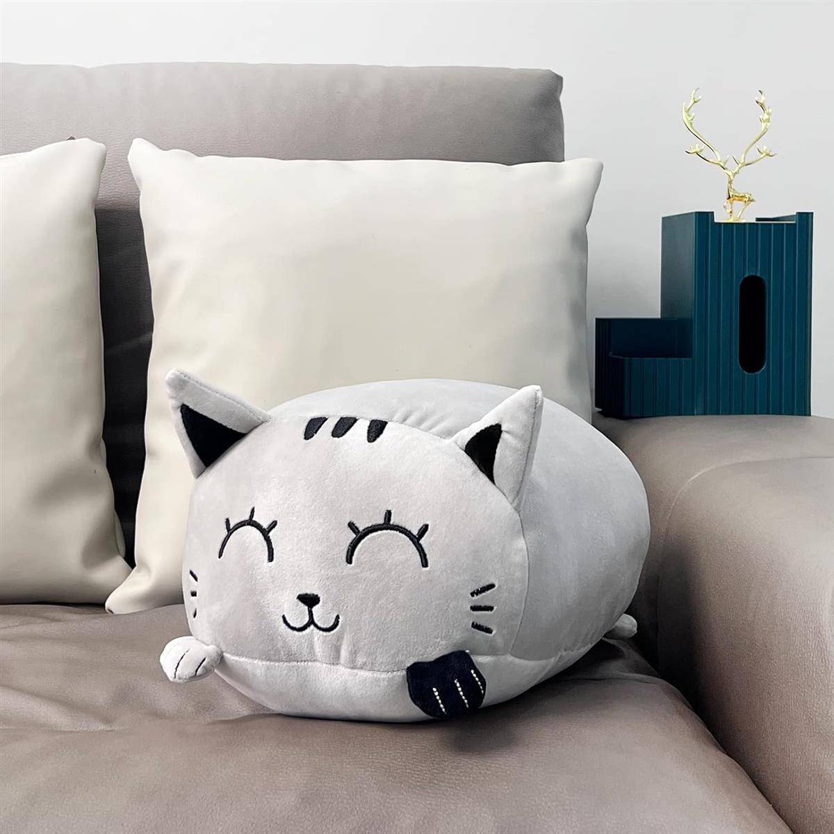 Plüschkissen in Katzenform auf einem beigen Sofa mit weißen Kissen, neben einer kleinen blauen Dekokiste und einer goldenen Verzierung.
