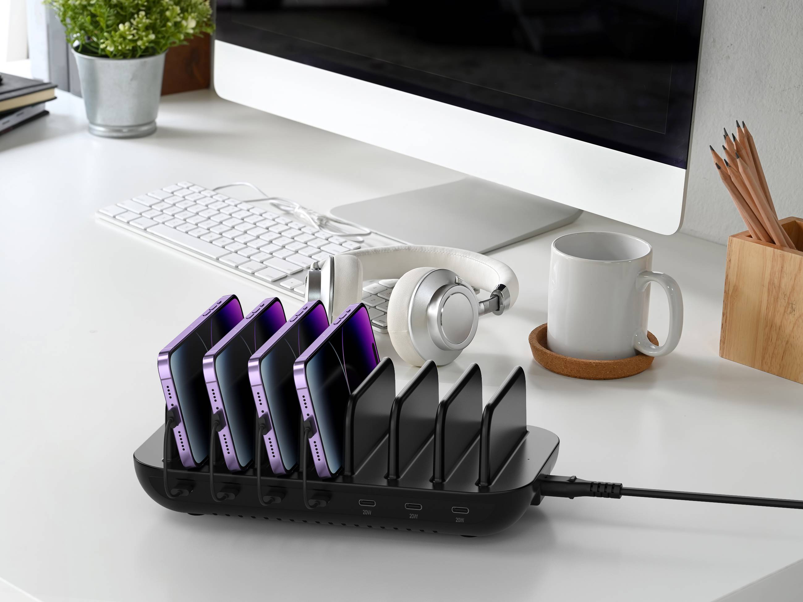 Sechs Smartphones laden in einer schwarzen Multi-Geräte-Ladestation auf einem Schreibtisch neben einem Computer, einer Tastatur, Kopfhörern und einer Kaffeetasse.