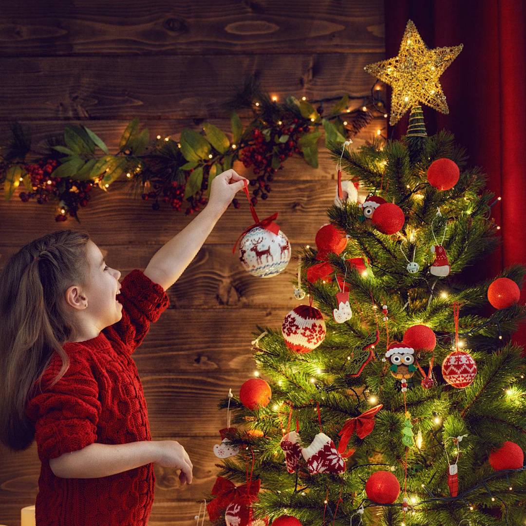 Ein junges Mädchen in einem roten Pullover hängt eine Weihnachtskugel an einem dekorierten Weihnachtsbaum auf, der mit Lichtern, roten Ornamenten und einer leuchtenden Sternenspitze geschmückt ist.