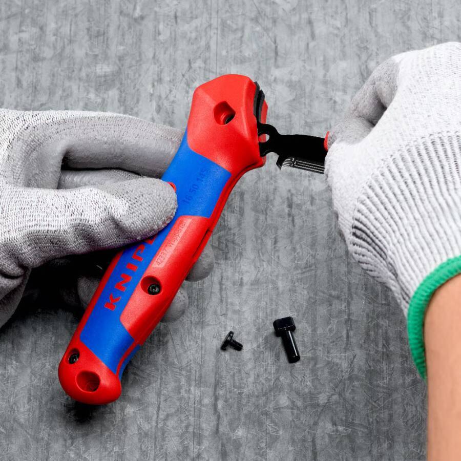 Behandschuhte Hände verwenden einen Schraubendreher, um eine Schraube aus einem Werkzeug mit rot-blauem Griff auf einer grauen Oberfläche zu entfernen, mit zwei kleinen Schrauben in der Nähe.