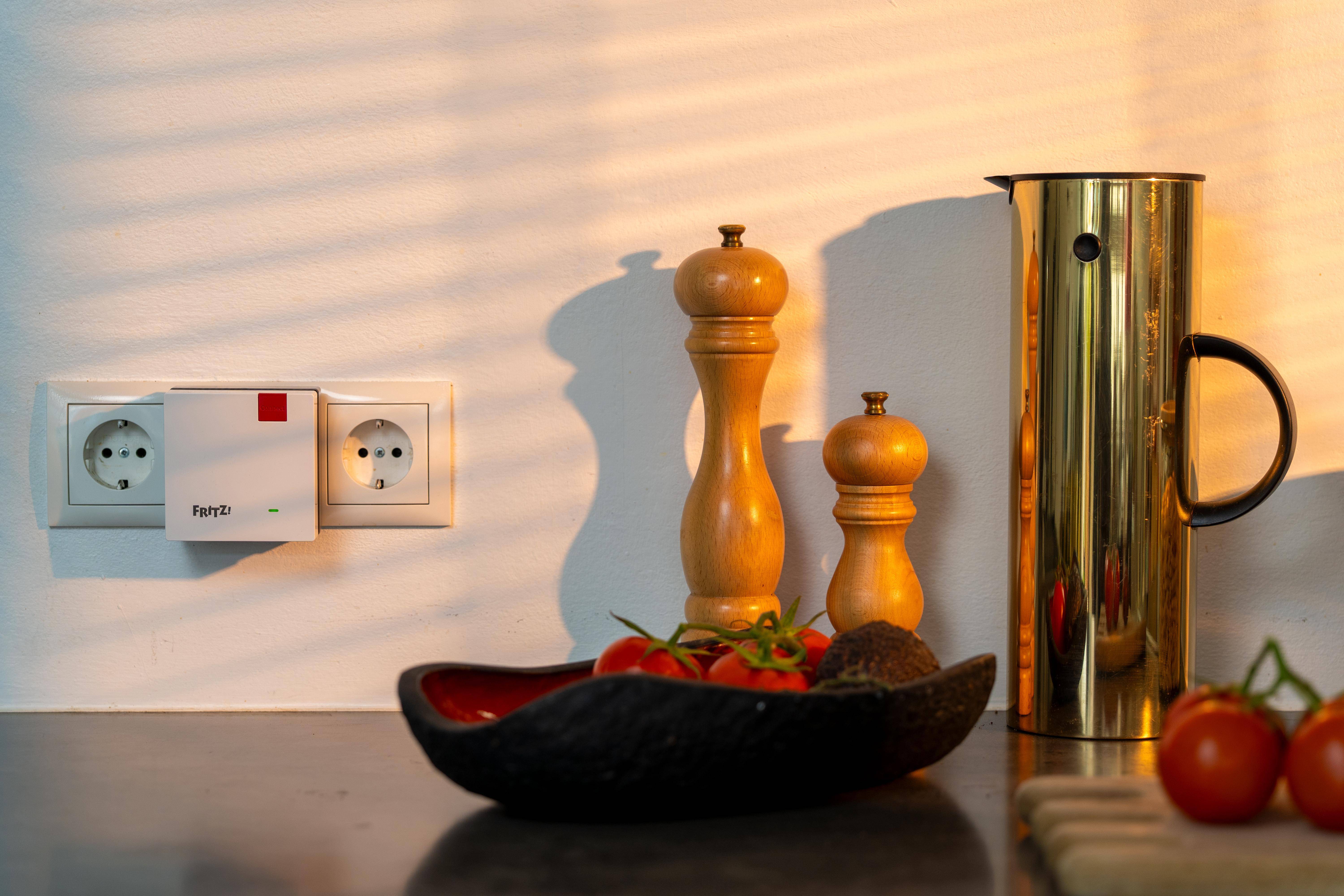 Küchenarbeitsplatte mit Pfeffermühlen, einem Metallkrug, Tomaten und einer Obstschale neben einer Wandsteckdose und einem angeschlossenen Gerät.