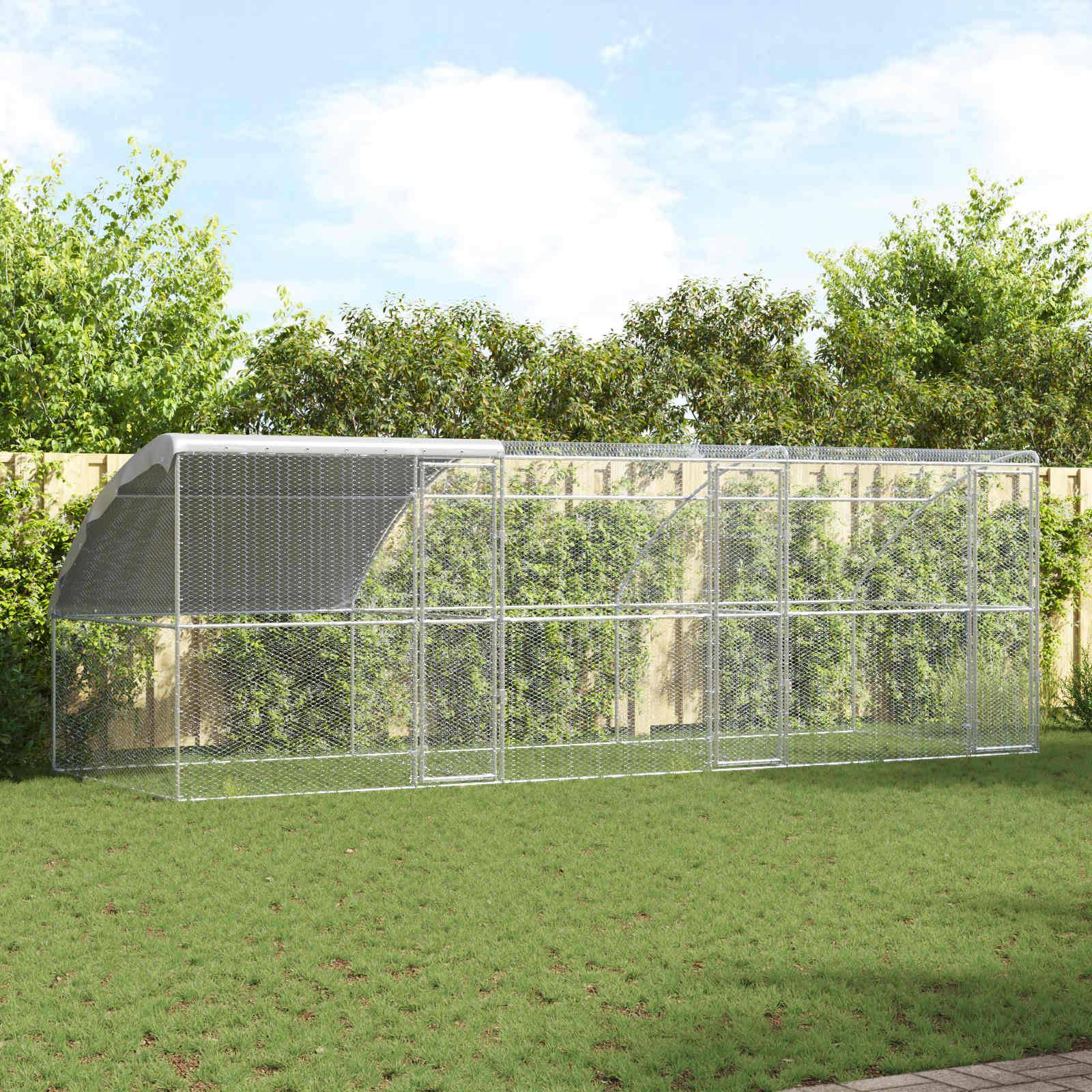 Ein großer Hühnerstall aus Metalldraht steht auf Gras, umgeben von Büschen und einem Zaun, unter einem klaren blauen Himmel.