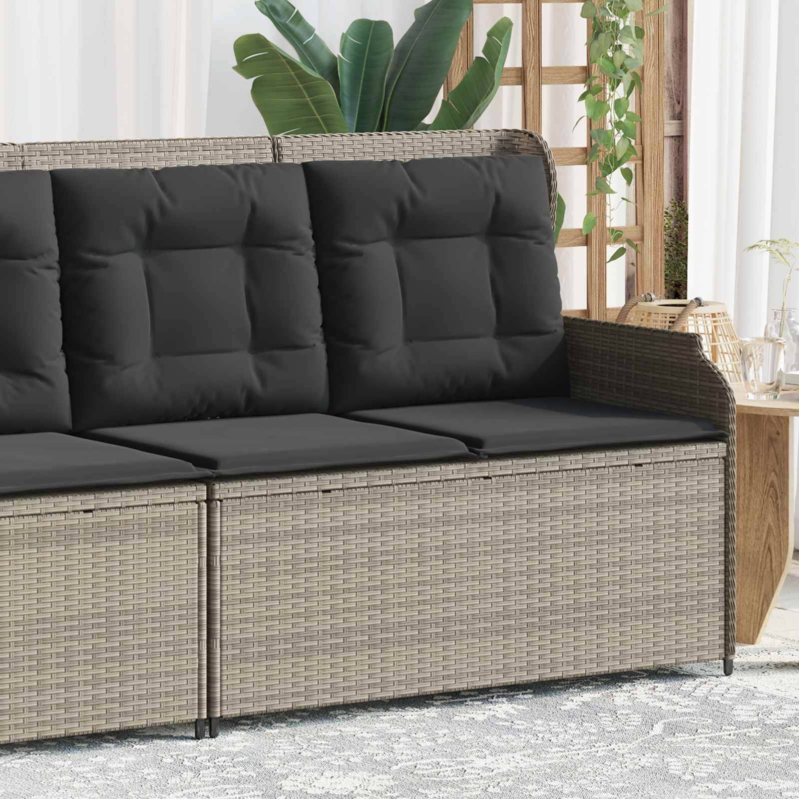 Rattansofa mit dunklen Kissen auf einer Terrasse, flankiert von grünen Pflanzen, neben einem kleinen Holzseitentisch mit einer Glaskanne und Gläsern.