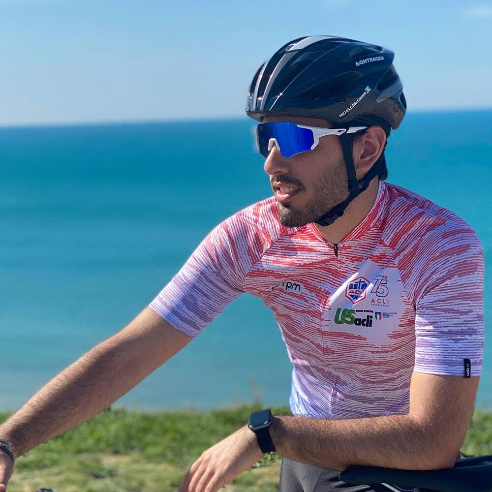 Ein Radfahrer mit Helm und Sonnenbrille fährt an einem sonnigen Tag, trägt ein rot-weißes Trikot mit verschiedenen Logos, in der Nähe eines Grünflächenbereichs.