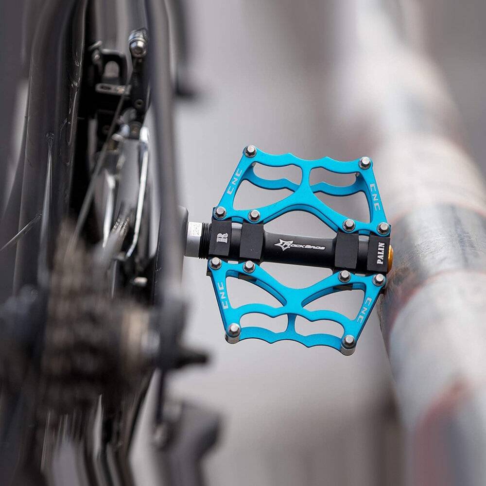 Eine Nahaufnahme eines Fahrradpedals, das an einem Fahrradrahmen befestigt ist, mit einem einzigartigen blauen Design und sichtbarem Markentext, positioniert in der Nähe eines Metallgeländers.