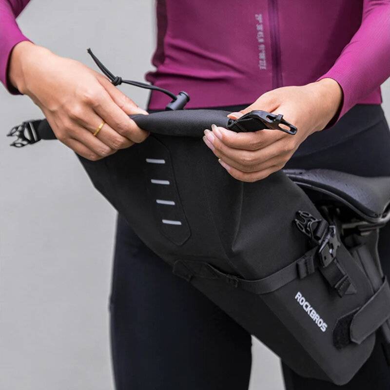 Eine Person in einem lila Radsport-Trikot befestigt eine schwarze Bikepacking-Tasche an ihrem Fahrrad und konzentriert sich dabei auf den Befestigungsprozess.