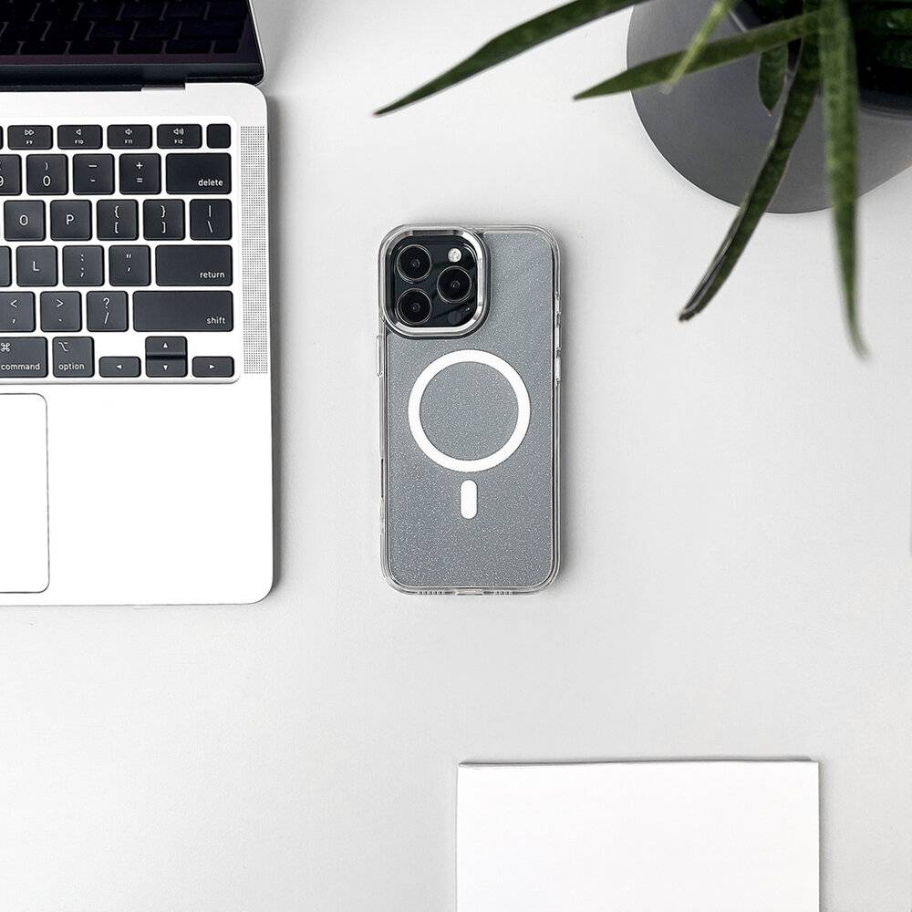 Ein Smartphone mit einer durchsichtigen Hülle und kreisförmigem Design liegt neben einem geöffneten silbernen Laptop auf einem weißen Schreibtisch, mit einer Pflanze in der Nähe.