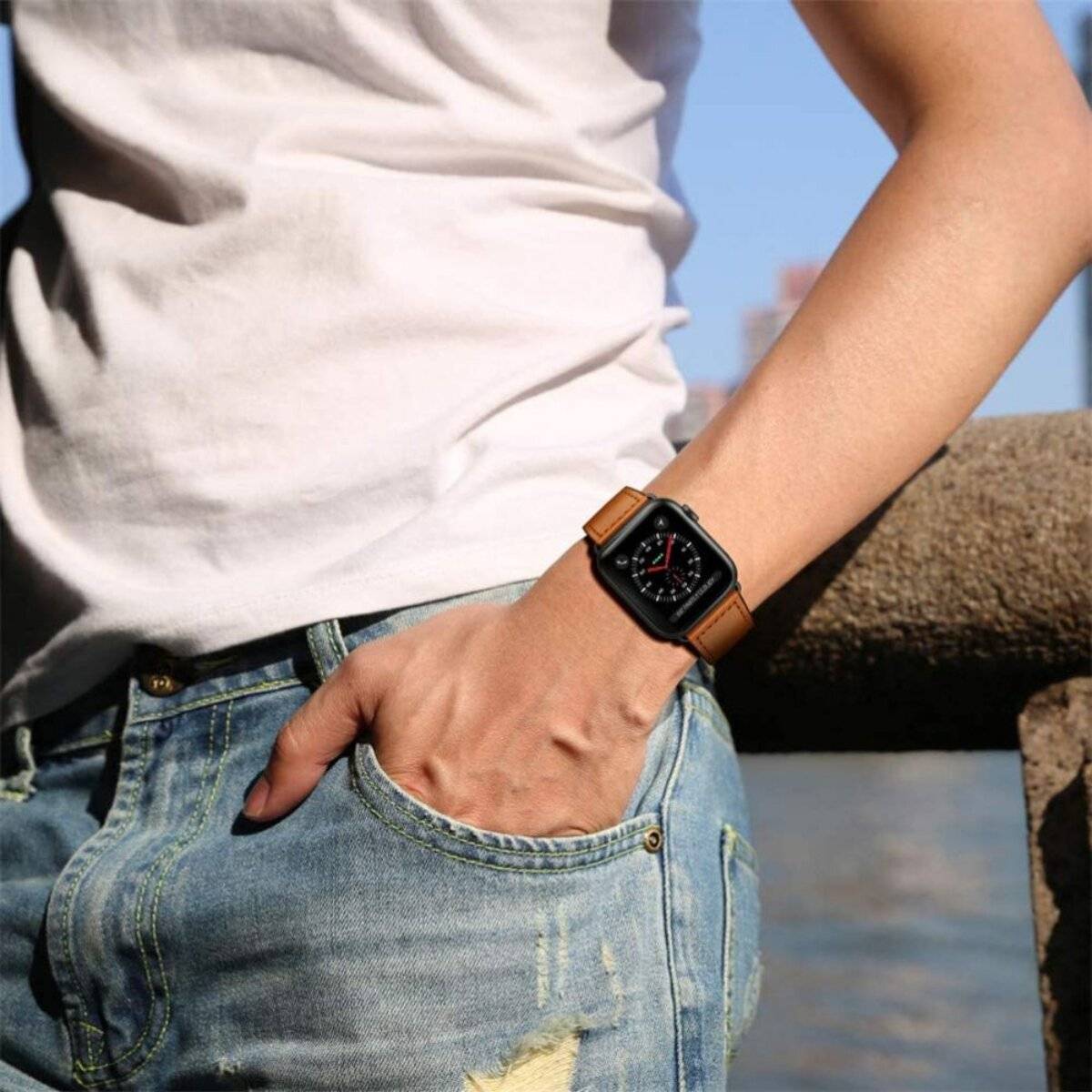 Eine Person trägt ein weißes T-Shirt und Jeans mit einer Hand in der Tasche. Sie haben eine Smartwatch am Handgelenk. Der Hintergrund ist unscharf.