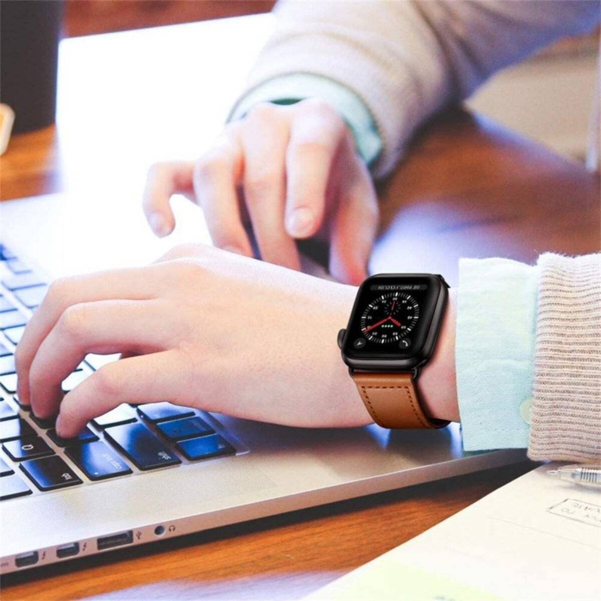 Eine Person tippt auf einem Laptop und trägt eine Smartwatch mit einem Lederarmband. Das Zifferblatt der Uhr zeigt eine analoge Uhrzeit mit einem roten Sekundenzeiger an.