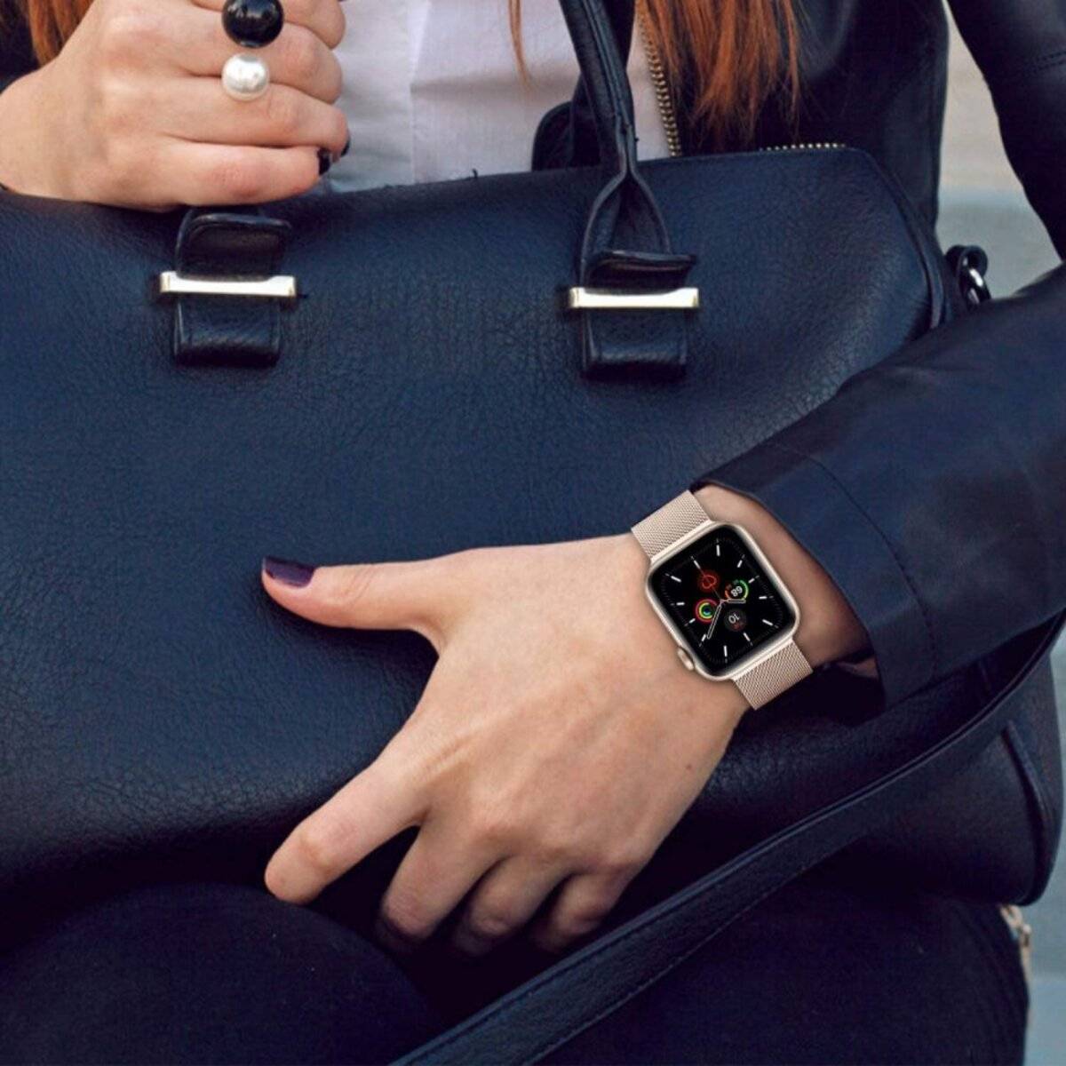 Eine Person sitzt und hält eine schwarze Handtasche, trägt eine Smartwatch mit einem farbigen Display, auf dem App-Symbole angezeigt werden, und trägt eine schwarze Jacke.