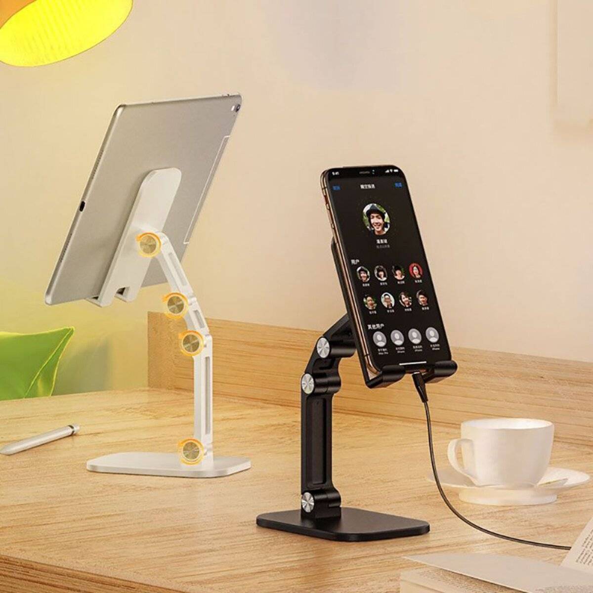 Zwei verstellbare Gerätehalter stehen auf einem Holzschreibtisch: einer hält ein Tablet unter einer Lampe, und der andere hält ein Smartphone mit einem Ladekabel in der Nähe.