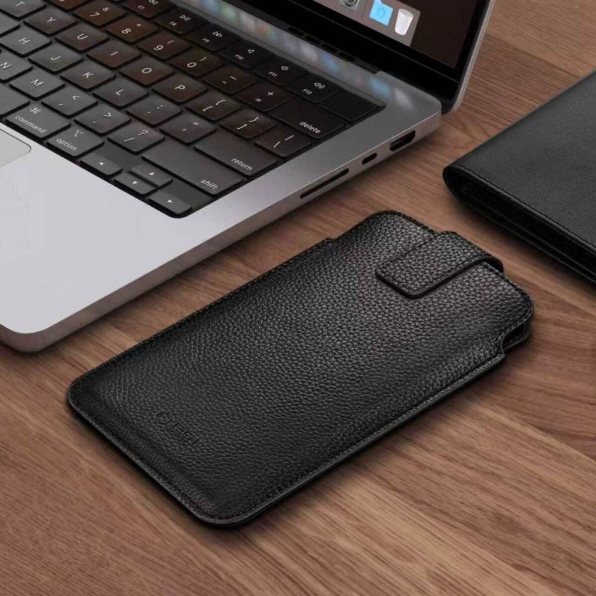 Ein schwarzes Leder-Smartphone-Case liegt auf einem Holzschreibtisch neben einem aufgeklappten Laptop und zeigt einen Teil der Tastatur und des Bildschirms.