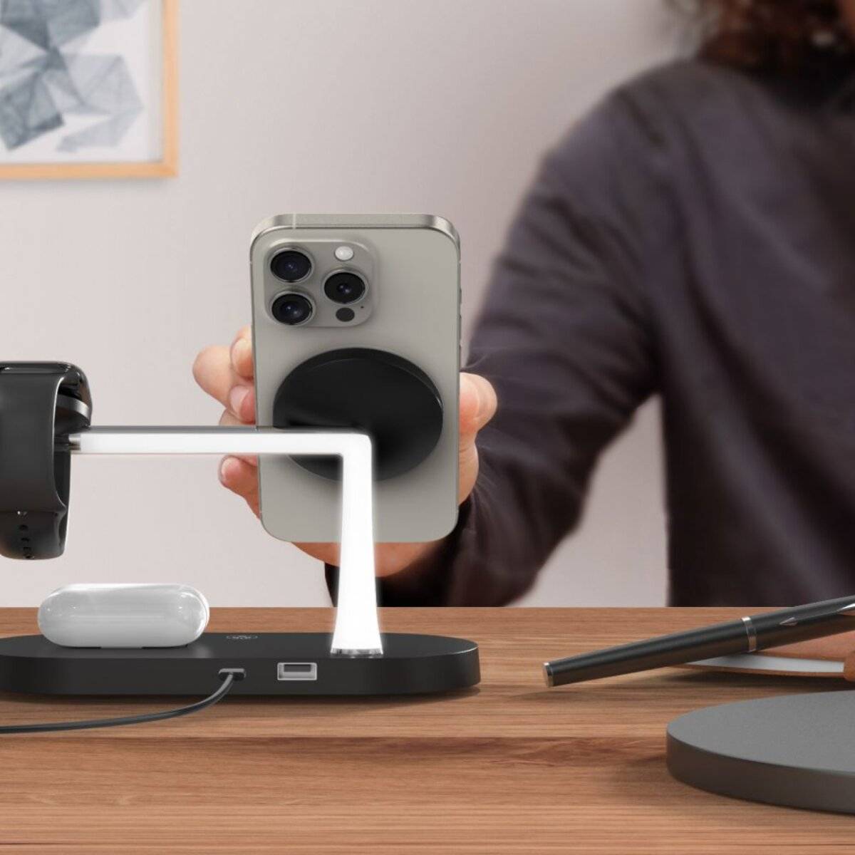 Eine Person legt ein Smartphone auf eine kabellose Ladestation, die auch eine Smartwatch und Ohrhörer auf einem Schreibtisch-Setup hält.
