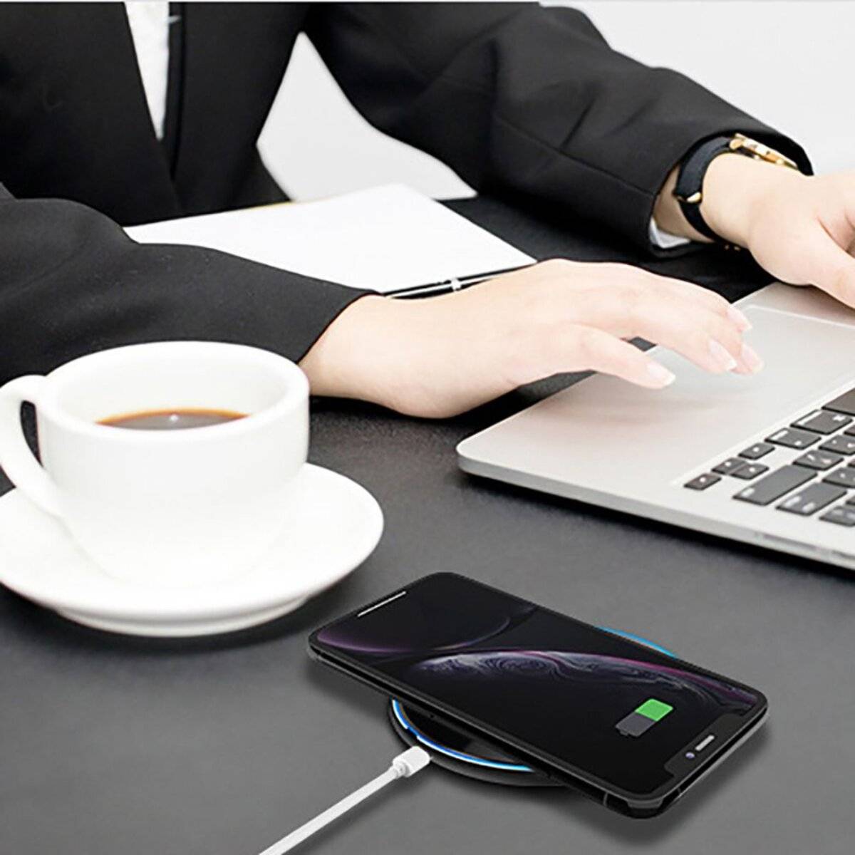 Eine Person, die an einem Laptop an einem Schreibtisch arbeitet, mit einer weißen Kaffeetasse, einem drahtlos ladenden Smartphone in der Nähe und Papieren im Hintergrund.