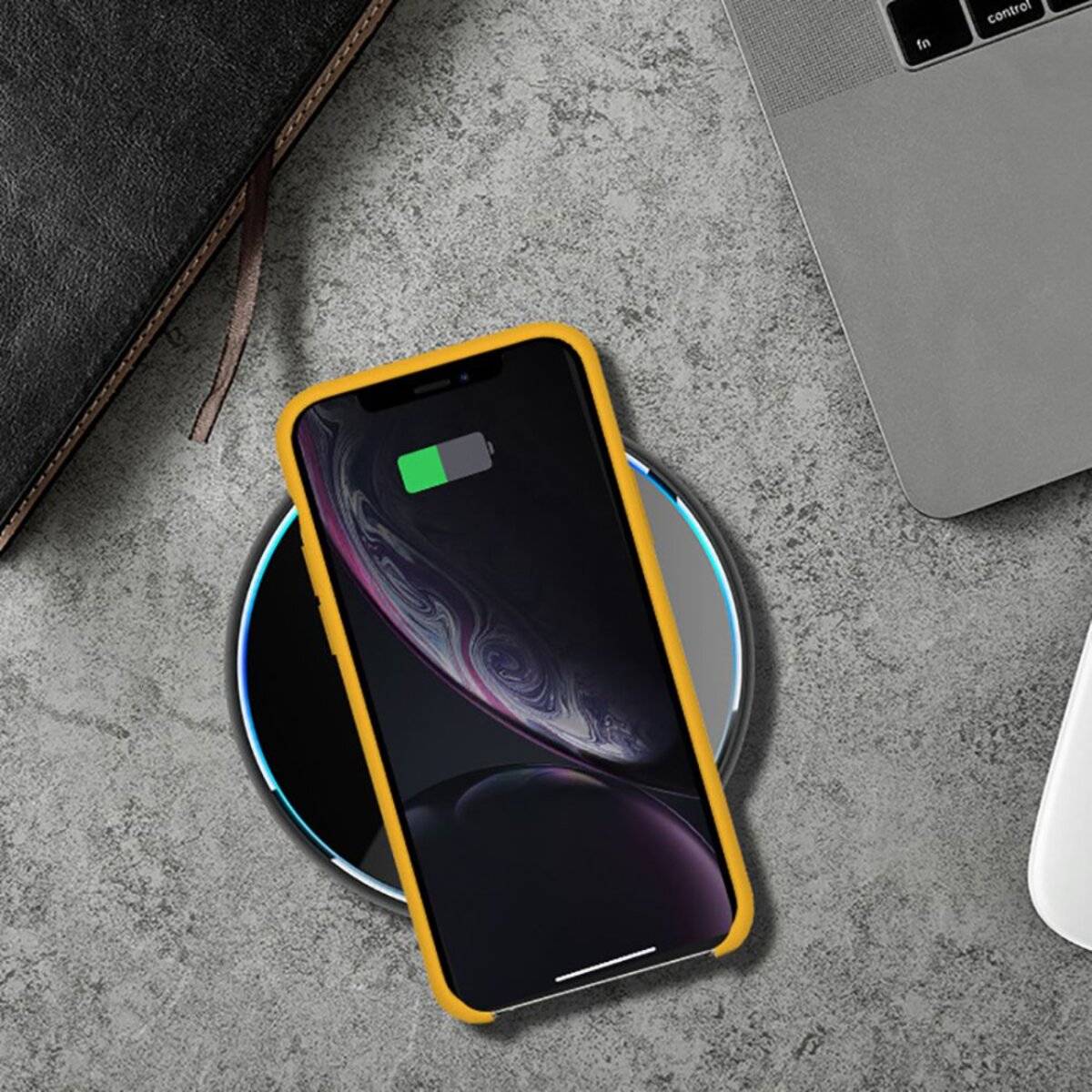 Ein Smartphone mit gelbem Case lädt sich drahtlos auf einer kreisförmigen Matte auf, die neben einem Laptop und einem Notizbuch auf einer grauen Oberfläche steht.