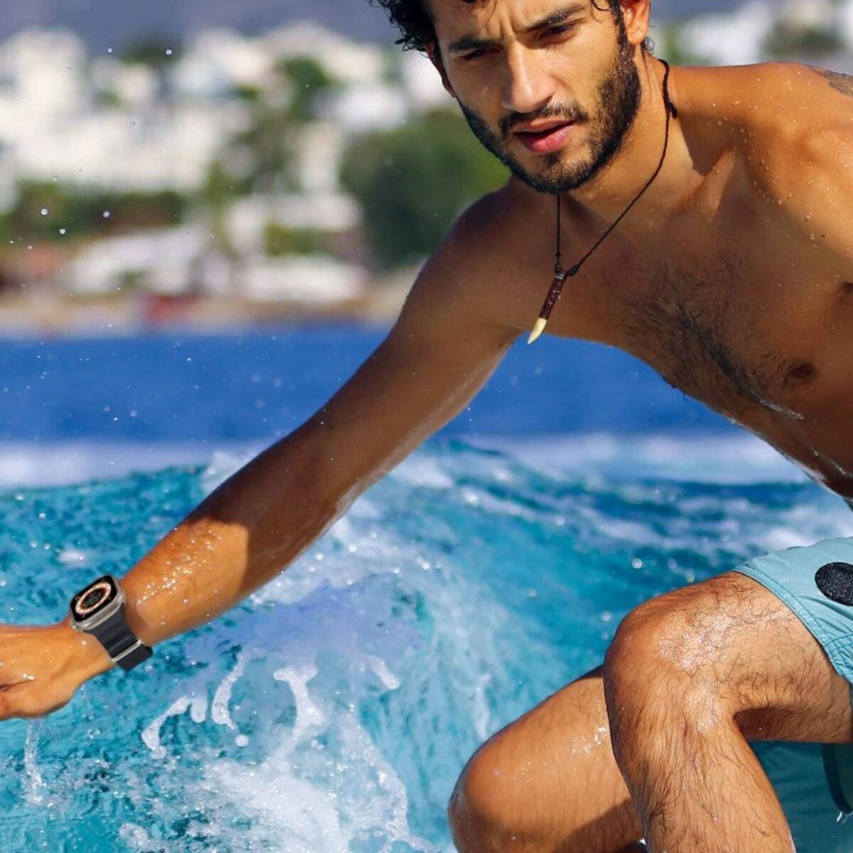 Ein Mann balanciert auf einem blauen Surfbrett und reitet eine Welle, dabei trägt er eine schwarze Armbanduhr und eine Halskette. Im unscharfen Hintergrund eine Küstenstadt und Berge.