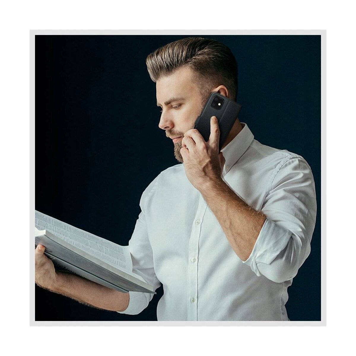 Ein Mann in einem weißen Hemd, der ein Buch hält und am Telefon spricht, wirkt konzentriert und engagiert im Gespräch vor einem dunklen Hintergrund.
