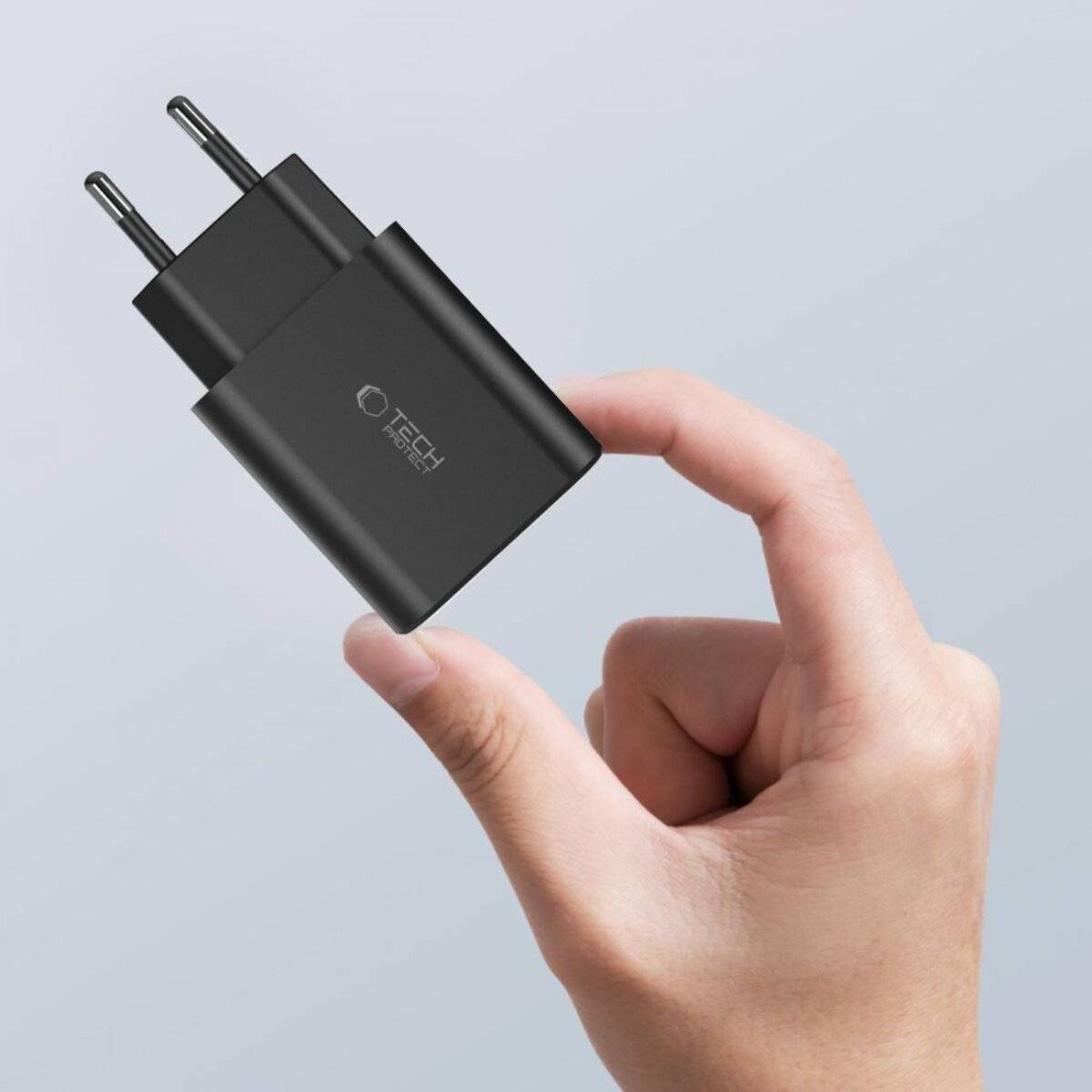 Eine Hand hält ein schwarzes Telefon-Ladegerät der Marke 'Tech' mit zwei Metallstiften, vor einem einfarbigen Hintergrund.