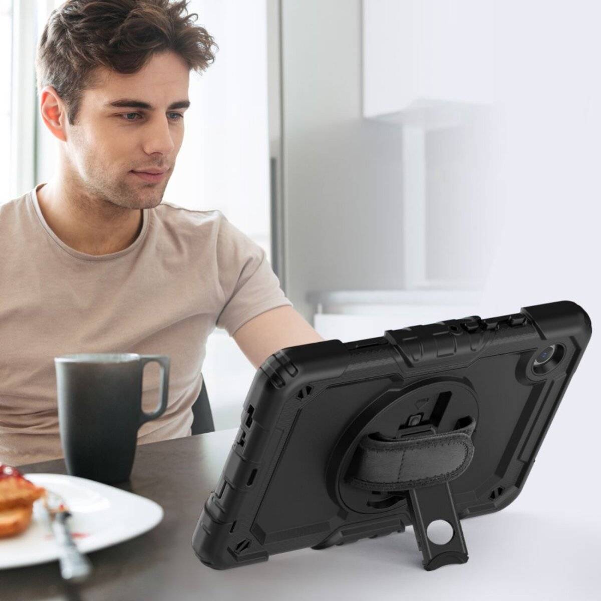 Ein Mann mit braunem Haar schaut auf ein Tablet auf einem Tisch, neben ihm liegen ein Teller mit Gebäck und eine Tasse in einem hellen Raum.
