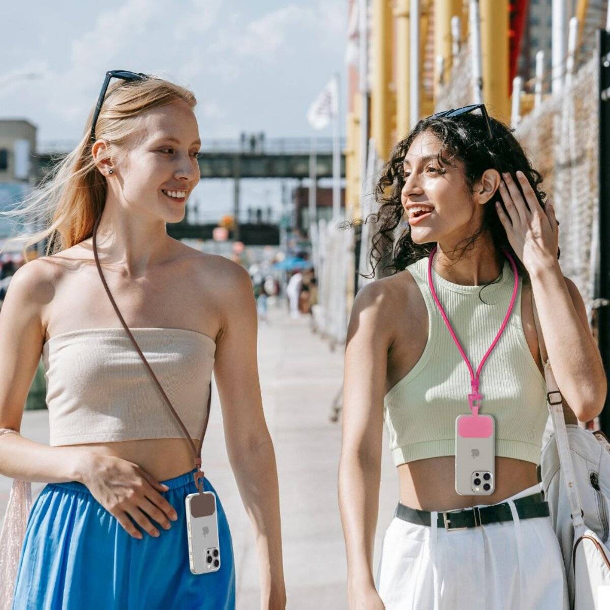 Zwei Frauen gehen an einem sonnigen Tag draußen spazieren, lächeln und tragen Sommerkleider mit Smartphones, die um ihre Hälse hängen.