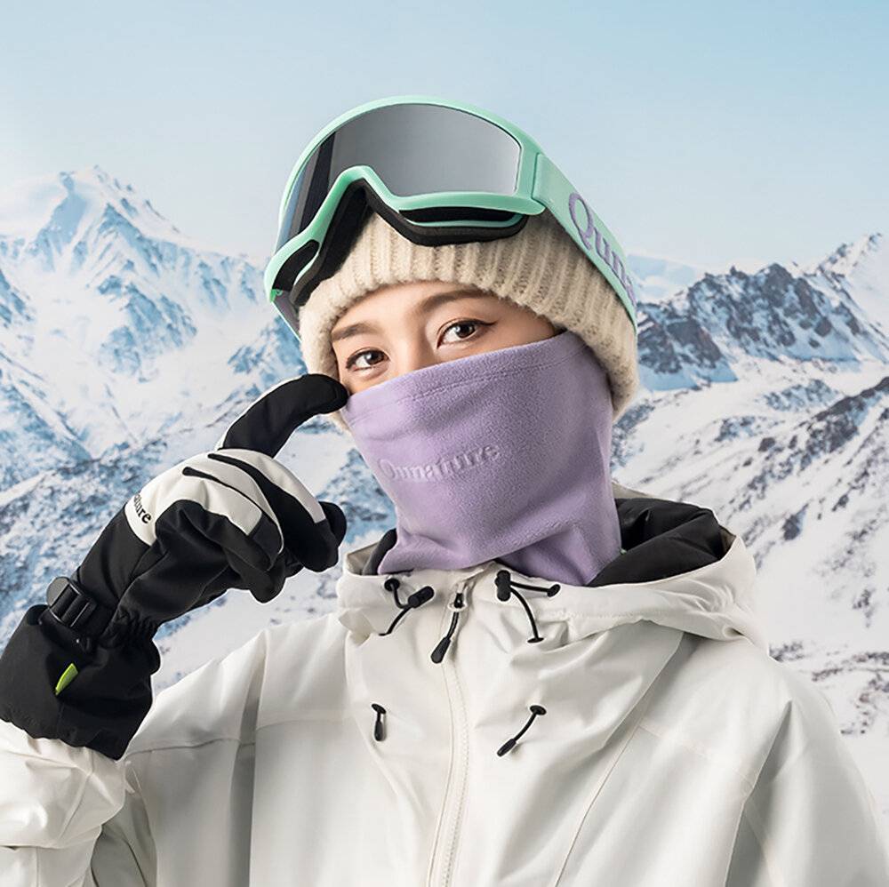 Eine Person in Schneeausrüstung, einschließlich Schutzbrille und Gesichtsmaske, steht vor einer verschneiten Berglandschaft und signalisiert mit einer Handbewegung 