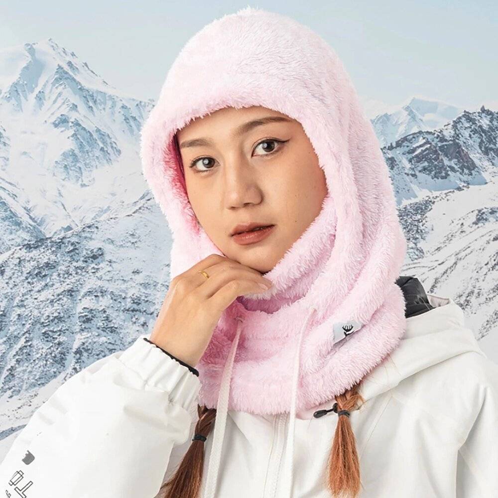 Eine Person trägt einen rosa, flauschigen Kapuzenhalstuch und steht vor einer verschneiten Bergkulisse, während sie direkt in die Kamera blickt.