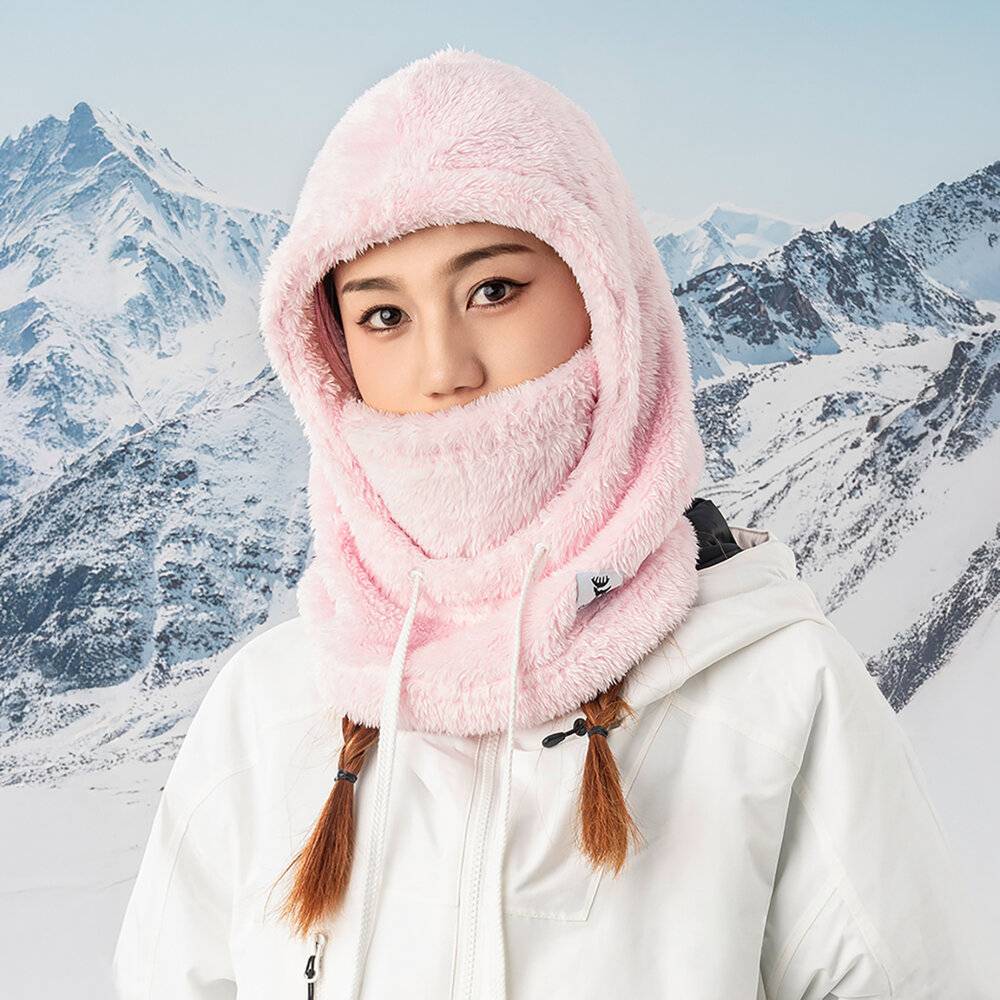 Eine Person trägt eine rosa flauschige Kapuze und Gesichtsbedeckung und steht vor einer schneebedeckten Bergkulisse, gekleidet in eine weiße Winterjacke.
